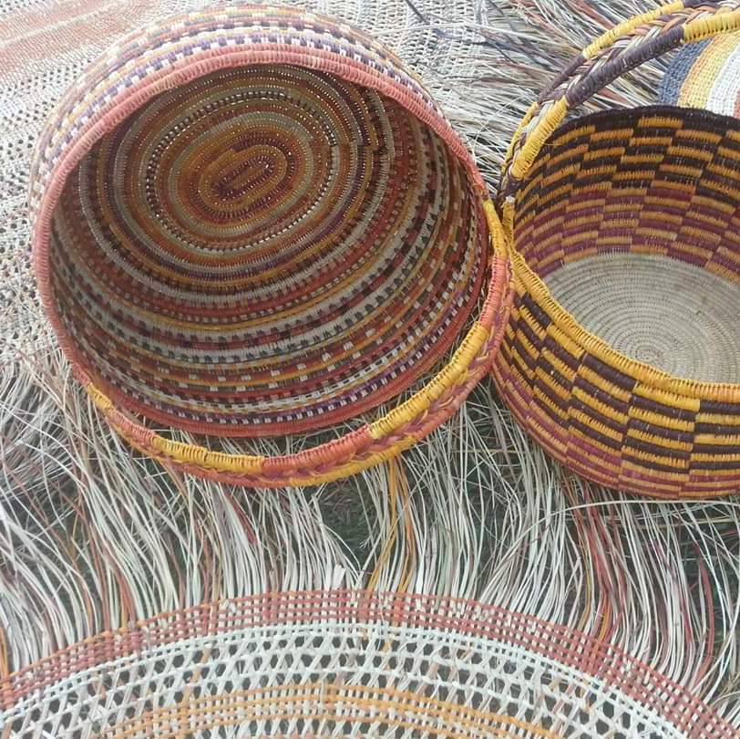 Two intricately woven Indigenous baskets, one showing a colorful interior and the other checkered, rest on a fringed fiber mat.